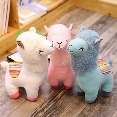 three stuffed llamas sitting next to each other on a wooden floor in front of a bookshelf