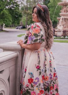 a woman in a floral dress leaning against a wall with her hands on her hips