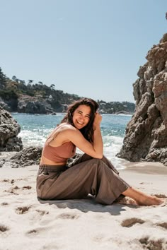 a woman sitting on top of a sandy beach next to the ocean with rocks in the background