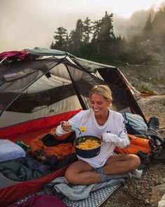 a woman sitting in front of a tent eating out of a bowl on the ground