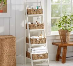 a white shelf with baskets and towels on it in front of a window next to a wooden bench