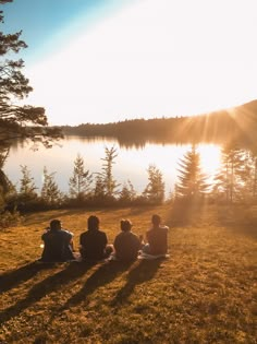 three people are sitting on the grass watching the sun go down over a body of water