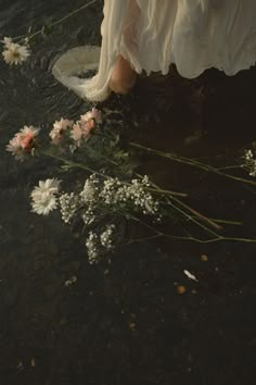 a woman in white dress sitting on the edge of a body of water next to flowers