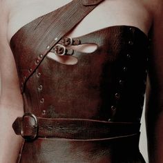 a woman wearing a brown leather dress with straps and buttons on her chest, in front of a white background