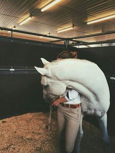 a woman is hugging a white horse in a stable with hay on the floor and ceiling