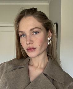 a woman with blue eyes wearing a brown coat and earrings on her left ear, standing in front of a white door