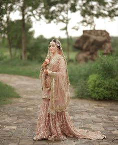 a woman in a pink dress standing on a brick path with trees and bushes behind her