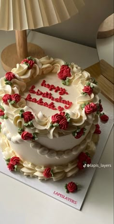 a cake with white frosting and red roses on it sitting on a table next to a lamp