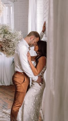 a bride and groom kissing in front of a white brick wall with flowers on it