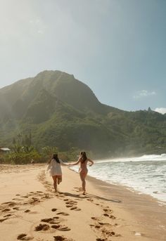 two women walking on the beach with footprints in the sand and mountains in the background