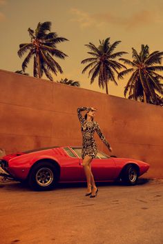 a woman standing next to a red sports car in front of palm trees and a wall