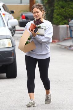 a woman is walking down the street with plants in her hand and holding a brown paper bag