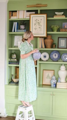 a woman standing on a stool in front of a green bookcase with plates and vases