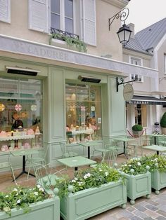 the outside of a store with green tables and chairs in front of it, surrounded by potted plants