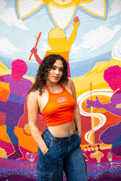 woman modeling upper park's club graphic baby rib halter top in orange in downtown chico, california standing in front of a mural. how to style halter top. cropped halter tank outfit. Fitted Tank Halter Top For Spring, Fitted Halter Top Tank For Spring, Stretch Halter Top With Tank Straps For Summer, Fitted Spring Halter Top With Tank Straps, Casual Ribbed Racerback Halter Top, Trendy Ribbed Tank Halter Top, Fitted Ribbed Halter Neck Top, Ribbed Fitted Halter Neck Top, Casual Cropped Halter Top With Stretch