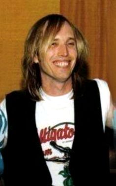 a man with long hair smiling and wearing a white t - shirt, black vest and blue jeans