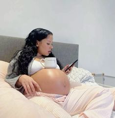 a pregnant woman laying in bed with her cell phone and holding a cup while looking at her stomach