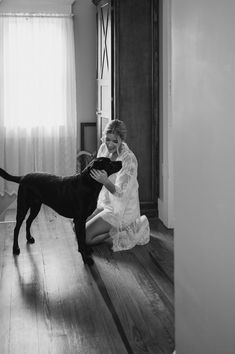 a woman kneeling down next to a dog on a hard wood floor in front of a window