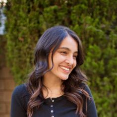 a woman smiling and wearing a black shirt in front of some green bushes with her hands on her hips