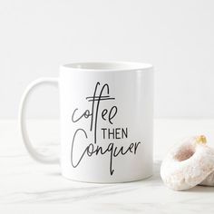 a white coffee mug with the words coffee then conquer written on it next to a donut