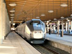 a silver train traveling down tracks next to a loading platform with lights on it's sides