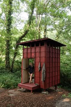 a woman standing in the doorway of a red outhouse surrounded by trees and foliage