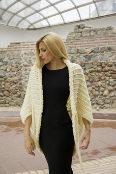 a woman wearing a black dress and a white crochet cardigan is standing in front of a stone wall