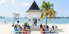 a wedding ceremony on the beach with people sitting in chairs and one man standing at the alter