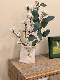 a vase with cotton flowers on a wooden table next to a framed photograph and a green frame