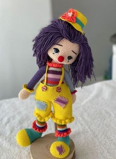 a crocheted doll with purple hair and yellow pants is standing on a wooden base