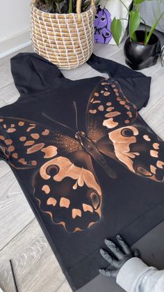 a black t - shirt with a butterfly on it sitting on the floor next to some plants