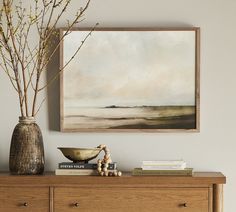 a painting hangs above a dresser with books and vases on it's side