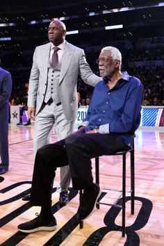 an older man in a suit and tie sitting on a chair next to a younger man