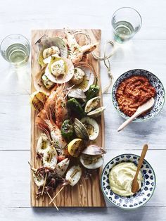 a wooden cutting board topped with seafood and veggies next to bowls of sauce