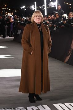 a woman in a brown coat and black dress standing on a gray carpet with cameras around her