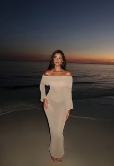 a woman standing on top of a beach next to the ocean at sunset with her hands in her pockets