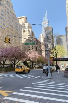 a yellow taxi driving down a street next to tall buildings in the city with pink flowers on trees