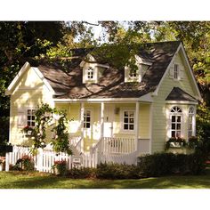 a small yellow house with white picket fence and flowers on the front porch, surrounded by trees