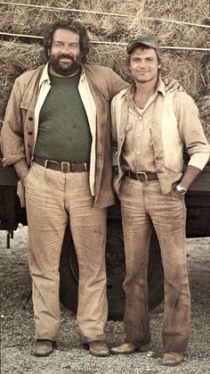 two men standing next to each other in front of a truck with hay on it