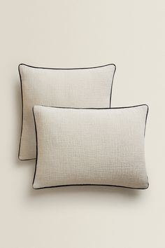 two white pillows with black piping on each side and one in the middle, against a beige wall