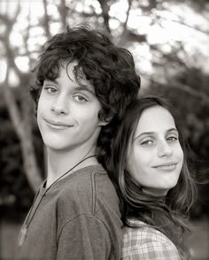 two young women standing next to each other in front of trees and bushes, one smiling at the camera