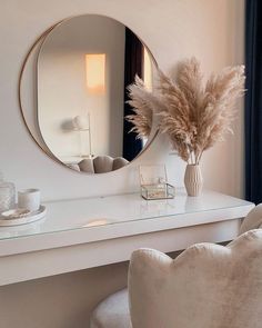 a white desk topped with a mirror and a vase filled with dry grass next to a chair