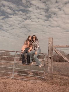 two young women sitting on top of a fence in front of a cloudy blue sky