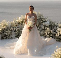 a woman in a wedding dress standing next to some bushes and flowers with the ocean in the background