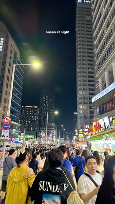 a crowd of people walking down a street at night in front of tall buildings and skyscrapers