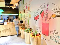 a man standing in front of a wall with fruit and drinks on it, next to two wooden planters filled with oranges