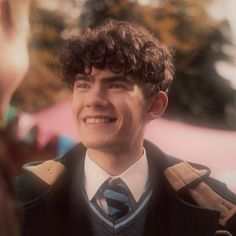 a young man with curly hair wearing a suit and tie smiling at someone in the background