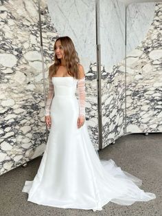 a woman standing in front of a marble wall wearing a white wedding dress with long sleeves