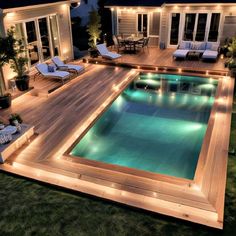 an outdoor swimming pool surrounded by decking and lights at night with lounge chairs around it