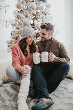 a man and woman sitting next to a christmas tree holding mugs